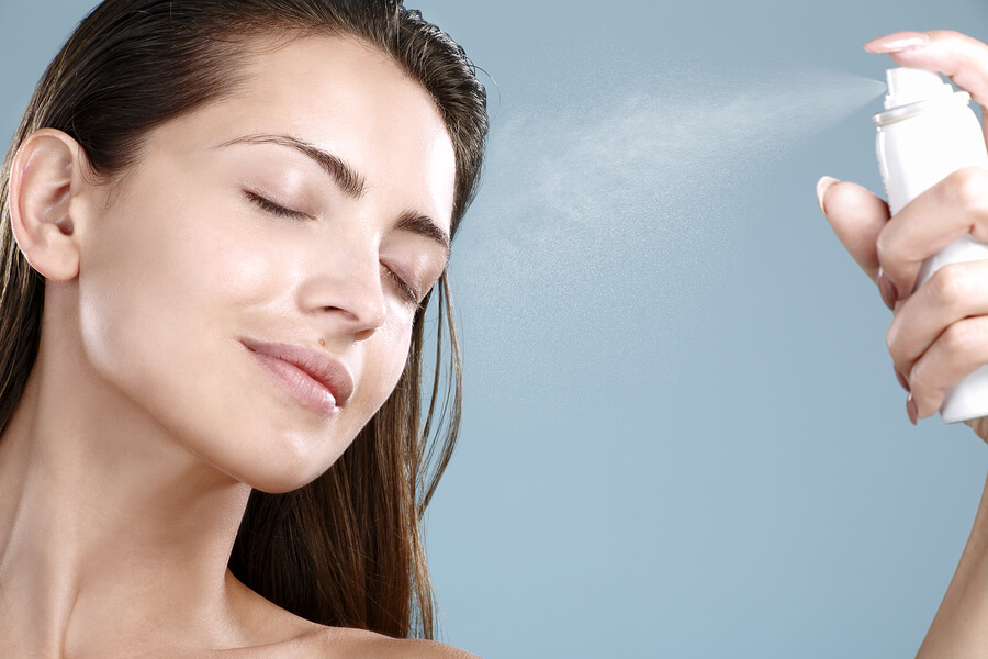 Beautiful woman applying spray water treatment on face on blue background